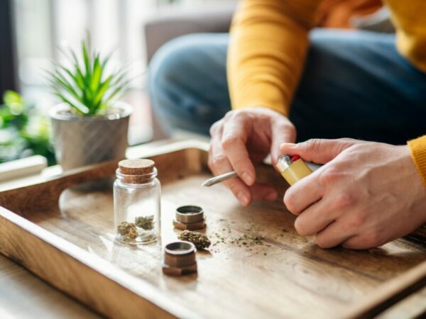 Ein Mann hält einen Joint in der Hand, dabei sieht man sein Gras und aufbewahrungsboxen.